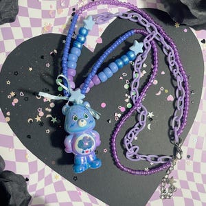 May include: A Care Bear necklace featuring a blue Care Bear pendant with a cloud design. The necklace has a beaded strand in shades of blue and purple, and a purple chain. The necklace is on a black heart-shaped surface.