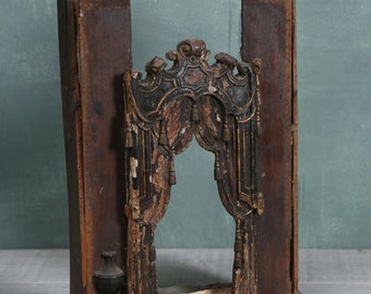 May include: A wooden frame with a carved, ornate design. The frame is painted black and gold and has a distressed look. The frame is missing a piece of wood on the bottom.