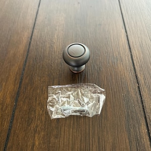 May include: A bronze-colored cabinet knob with a circular design, resting on a wooden surface. A small plastic bag containing a screw is also present. The knob has a rounded top and a short stem.