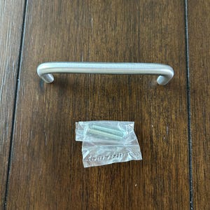 May include: A silver cabinet pull with a rounded rectangular shape. The pull is shown on a wood surface with two screws in a plastic bag.