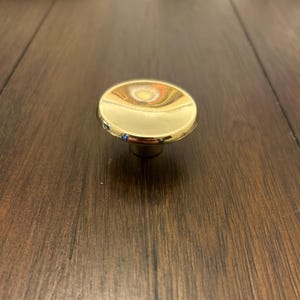 May include: A gold-colored cabinet knob with a rounded, flat top and a short, cylindrical base. The knob reflects light, creating a bright, shiny appearance. The hardware is designed for use on cabinets and drawers.