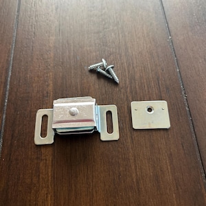 May include: A set of cabinet hardware components, including a silver magnetic catch, a small metal plate, and three silver screws. The hardware is designed for securing cabinet doors and drawers. The items are displayed on a dark brown wooden surface.