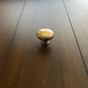 May include: A single, round cabinet knob with a brass finish. The knob has a smooth, reflective surface and a short, decorative base. The knob is centered on a dark brown wooden surface.