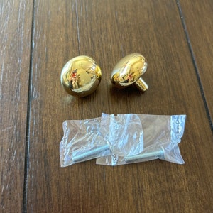 May include: Two gold-toned round cabinet knobs with a shiny finish and a screw attachment. Two silver screws are included in a clear plastic bag.