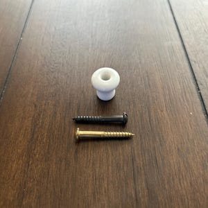 May include: A white ceramic cabinet knob with a black and a gold screw. The knob is round with a small, circular indentation. The screws are metal with a spiral design.