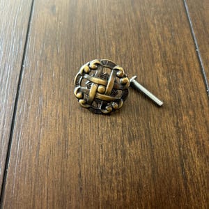 May include: A gold-toned metal cabinet knob with a woven design and a matching screw.