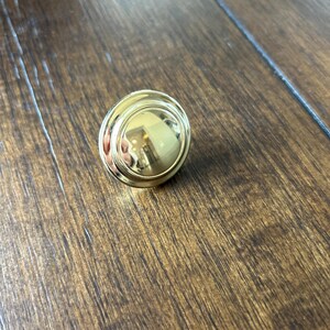 May include: A gold-toned round cabinet knob with a raised outer edge and a smooth, reflective surface.