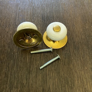 May include: Two white ceramic cabinet knobs with gold accents and mounting plates. The knobs are shown with two screws.