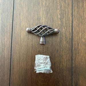 May include: A bronze-colored cabinet knob with a twisted wire design. The knob has a square base and two round knobs on either side. The knob is shown on a wood surface with two screws in a plastic bag.
