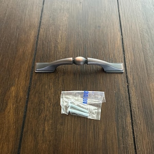 May include: A bronze-colored cabinet pull with a central oval detail and rectangular ends. Includes a small clear plastic bag containing screws. The hardware is designed for furniture or cabinetry.