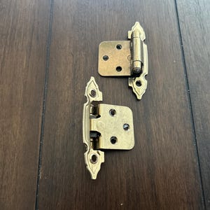 May include: Two brass-colored cabinet hinges with decorative edges are displayed on a dark wood surface. Each hinge has a rectangular plate with screw holes and a pivoting mechanism. The hinges are designed for cabinet doors.