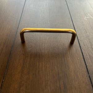 May include: A gold-colored cabinet pull handle with a rectangular shape. The handle is made of metal and has a smooth, shiny finish. The handle is set against a dark wood surface.