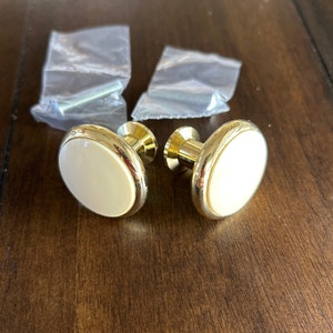 May include: Two gold cabinet knobs with white ceramic inserts. The knobs are round and have a smooth finish.