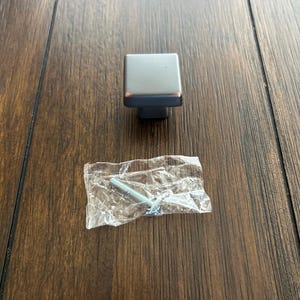 May include: A square, brushed nickel cabinet knob with a dark gray base and a screw in a clear plastic bag. The hardware is set against a dark brown wood grain surface.