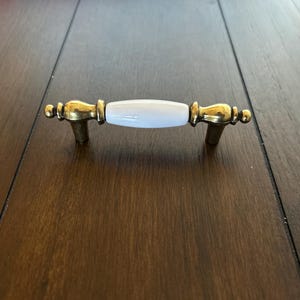May include: A cabinet pull with a white ceramic center and gold-toned metal ends. The pull has a classic design, with decorative detailing on the ends and a smooth, curved center. The hardware is designed for easy installation.