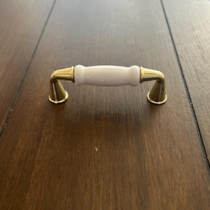 May include: A cabinet pull with a white ceramic center and gold-colored metal accents. The handle is a light lavender color. The hardware is designed for furniture and has a classic, elegant appearance.