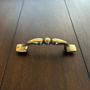 May include: A brass-toned cabinet pull with a curved handle and rectangular mounting plates. The handle has a central sphere and is set against a dark wood surface.