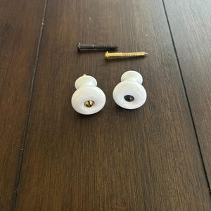 May include: Two white ceramic cabinet knobs with gold and black accents, alongside two screws. The knobs have a rounded shape and are designed for furniture or cabinetry. The screws are black and gold.