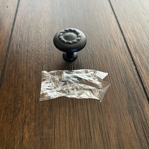 May include: A black cabinet knob with a decorative rope-like design around the top. The knob is shown with a small packet containing a screw, all resting on a wooden surface.