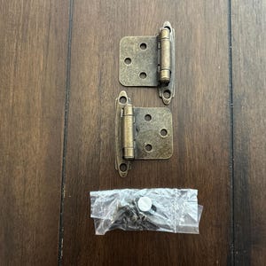 May include: Two antique brass cabinet hinges with mounting hardware. The hinges are rectangular with rounded edges and three screw holes. A small bag of screws and a white plastic piece are also included.