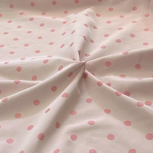 May include: A close-up of a white fabric with a pink polka dot pattern. The fabric is wrinkled and has a subtle texture.