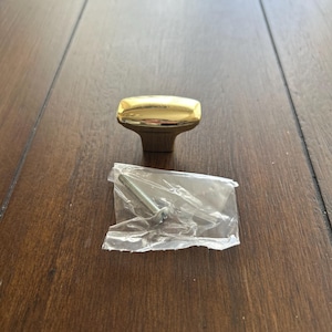 May include: A gold-colored cabinet knob with a rectangular top and a short stem. A screw is included in a small plastic bag. The knob is designed for furniture and home decor.