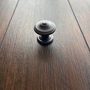 May include: A bronze-colored cabinet knob with a rounded top and a circular base. The knob has a classic design and is made of metal. The hardware is set against a dark wood grain background.