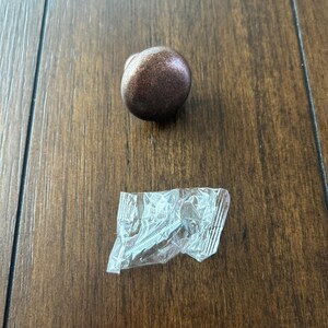 May include: A single, round, bronze-colored cabinet knob with a smooth, slightly textured surface. The knob is on a wooden surface.