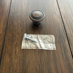 May include: A bronze-colored cabinet knob with a circular design, featuring a rounded top and a tiered base. A screw is included in a small, clear plastic bag. The knob is on a dark brown wooden surface.