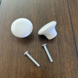 May include: Two white ceramic cabinet knobs and two silver screws are displayed on a brown wooden surface. One knob is round, and the other is a mushroom shape. The screws are straight with a threaded design.