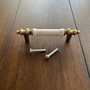 May include: A cabinet pull with a white ceramic center and gold-colored metal ends and posts. Two silver screws are included. The hardware is designed for furniture and home decor projects.