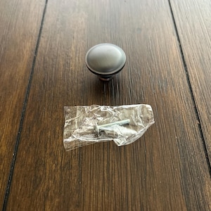 May include: A silver-colored cabinet knob with a dark brown base and a screw in a clear plastic bag. The knob is round and has a brushed metal finish. The hardware is designed for use on cabinets or drawers.