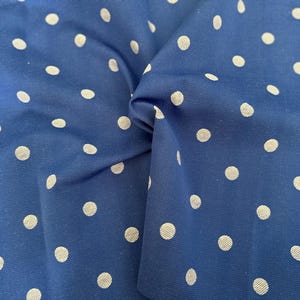 May include: Close-up of a blue fabric with a white polka dot pattern. The fabric is gathered and folded, showing the texture and design. The polka dots are evenly spaced and vary in size.