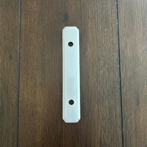 May include: A rectangular, off-white metal plate with two evenly spaced holes. The plate has a slightly raised border and is set against a dark brown wooden surface.