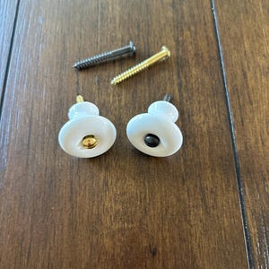 May include: Two white ceramic guitar strap buttons with gold and black accents. The buttons are accompanied by two screws, one black and one gold.