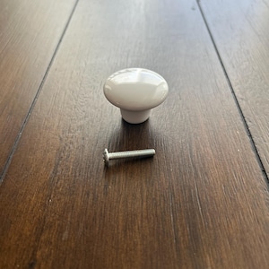 May include: A white ceramic cabinet knob with a rounded top and a short, cylindrical base. A silver screw is positioned next to the knob. The knob and screw are on a dark brown wooden surface.