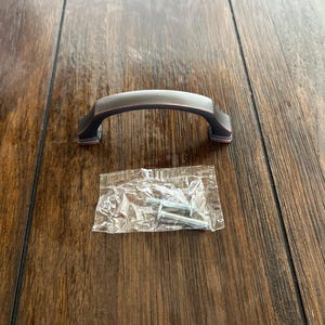 May include: A bronze-colored cabinet pull with a curved handle and square ends. A small clear plastic bag containing screws is next to the pull. The hardware is designed for use on cabinets and drawers.