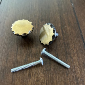 May include: Two gold-colored round cabinet knobs with a scalloped edge and black metal accents. Two silver screws are included.