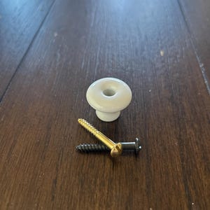 May include: A white ceramic cabinet knob with a gold screw and a black screw. The knob is round with a hole in the center. The screws are made of metal.