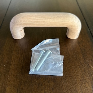 May include: A light brown wooden cabinet pull with a rounded shape. Two silver screws are included in a clear plastic bag.