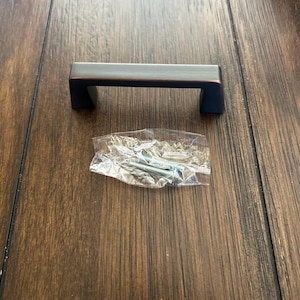 May include: A bronze-colored cabinet pull with a rectangular bar and two square feet. The pull is shown with a small plastic bag containing screws, likely for installation. The hardware is designed for furniture.