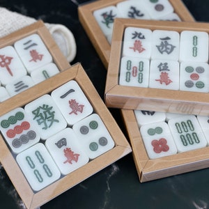 May include: A set of mahjong tiles in a wooden box. The tiles are white with black and red characters and symbols. The box is brown with a clear plastic lid.