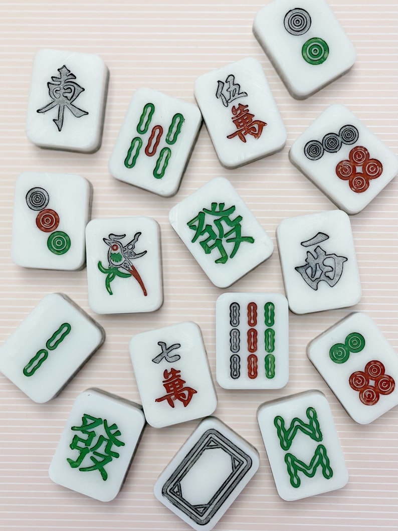 May include: A set of 14 white mahjong tiles with various Chinese characters and symbols in green, red, and black. The tiles are arranged in a random pattern on a pink and white striped background.