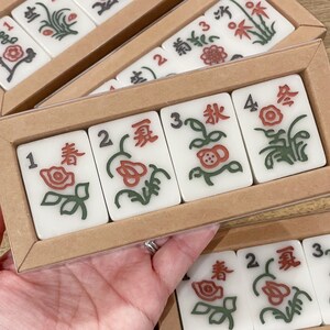 May include: A set of four white mahjong tiles with green and red floral designs and black numbers. The tiles are arranged in a row in a wooden box with a clear plastic lid.