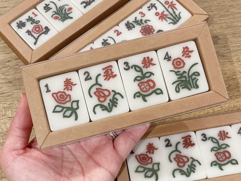 May include: A set of four white mahjong tiles with green and red floral designs and black numbers. The tiles are arranged in a row in a wooden box with a clear plastic lid.