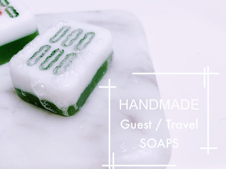 May include: Two green and white handmade soap bars shaped like mahjong tiles. The soap bars are on a white marble surface. The text "HANDMADE Guest / Travel SOAPS" is visible in the image.