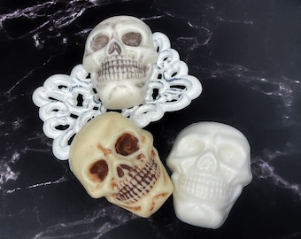 May include: Three white ceramic skull sculptures, two with painted details, on a black marble surface with a white decorative design.