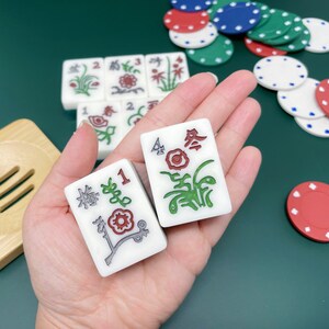 May include: Two white mahjong tiles with green and red floral designs. The tiles are numbered 1 and 4. The tiles are held in a hand against a green felt background.