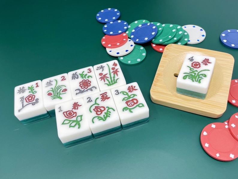 May include: A set of nine white mahjong tiles with green and red accents. The tiles feature Chinese characters and floral designs. The tiles are arranged in a row on a green surface. There are also several red and blue poker chips scattered around the tiles.