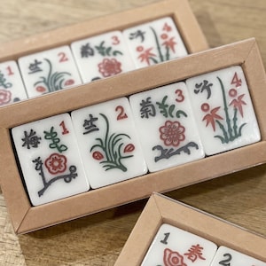 May include: A set of six white ceramic tiles with black and green floral designs and red flowers. The tiles are numbered 1, 2, 3, and 4. The tiles are arranged in a rectangular wooden box with a brown lid.